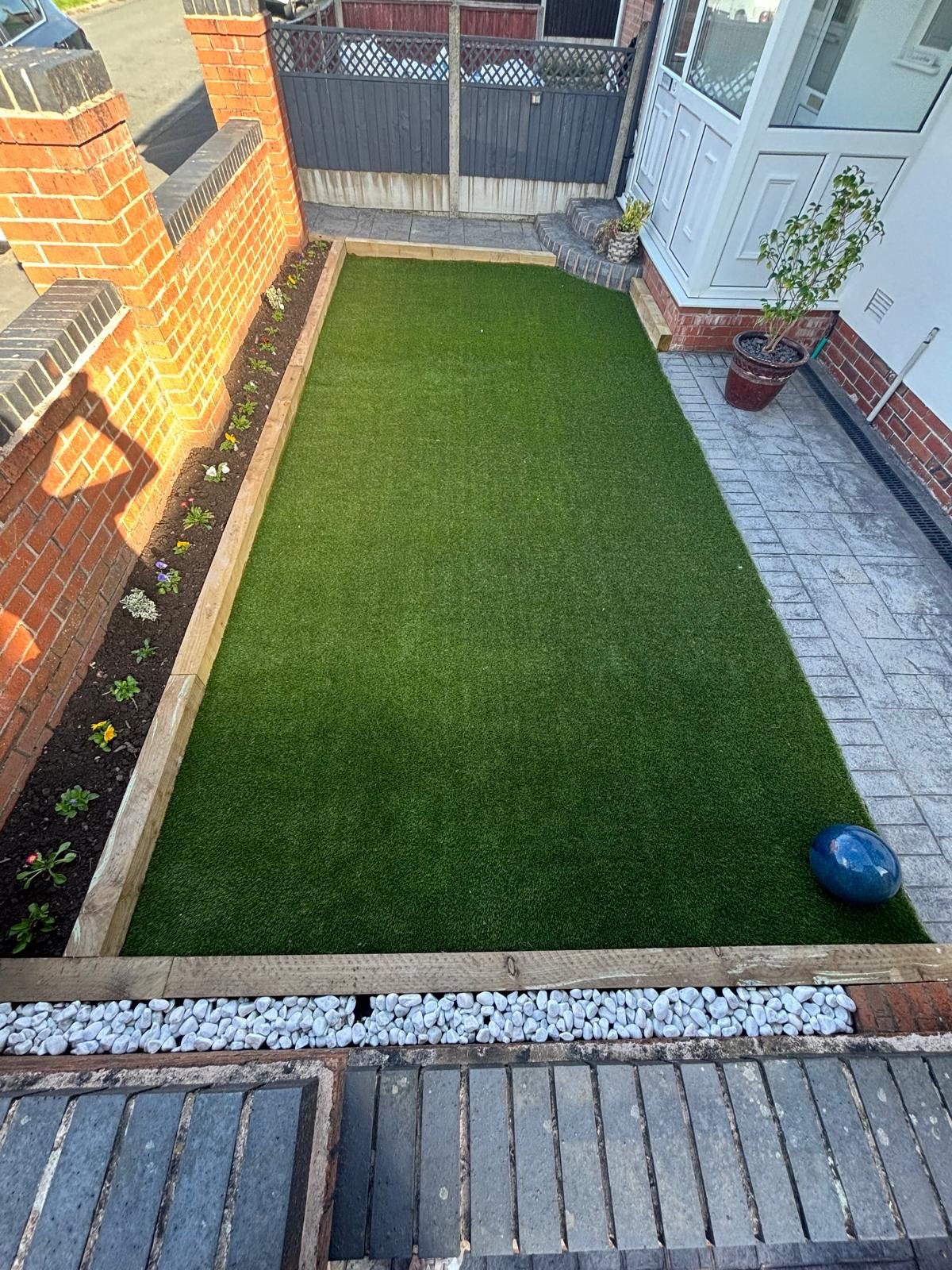 Finished front garden in sunshine: artificial lawn framed by railway sleepers, a planted border along the brick wall and a white pebble strip at the front