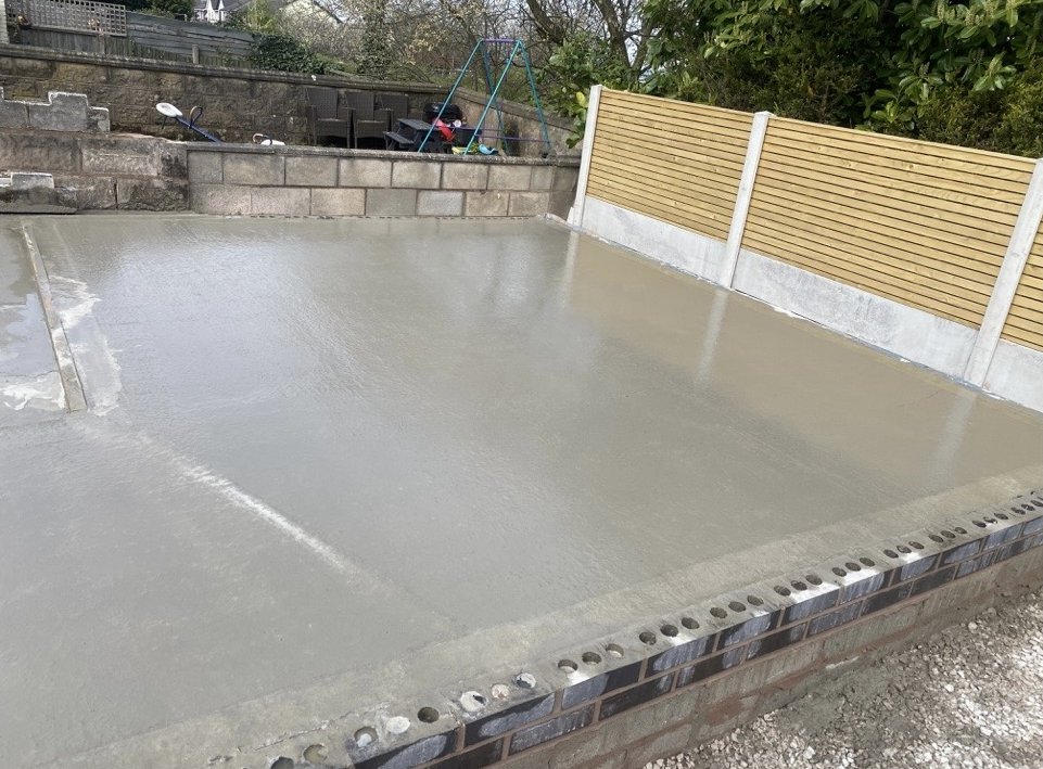 Freshly finished concrete base within a brick and block perimeter, slatted fence alongside